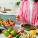 Woman in pink overshirt holds tomato in kitchen with fresh fruits and veggies.